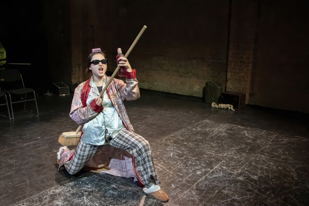 Actor wearing sunglasses, pajamas, and a curler in her hair holds a broom along with a water bottle filled with red liquid on stage in mid-performance.